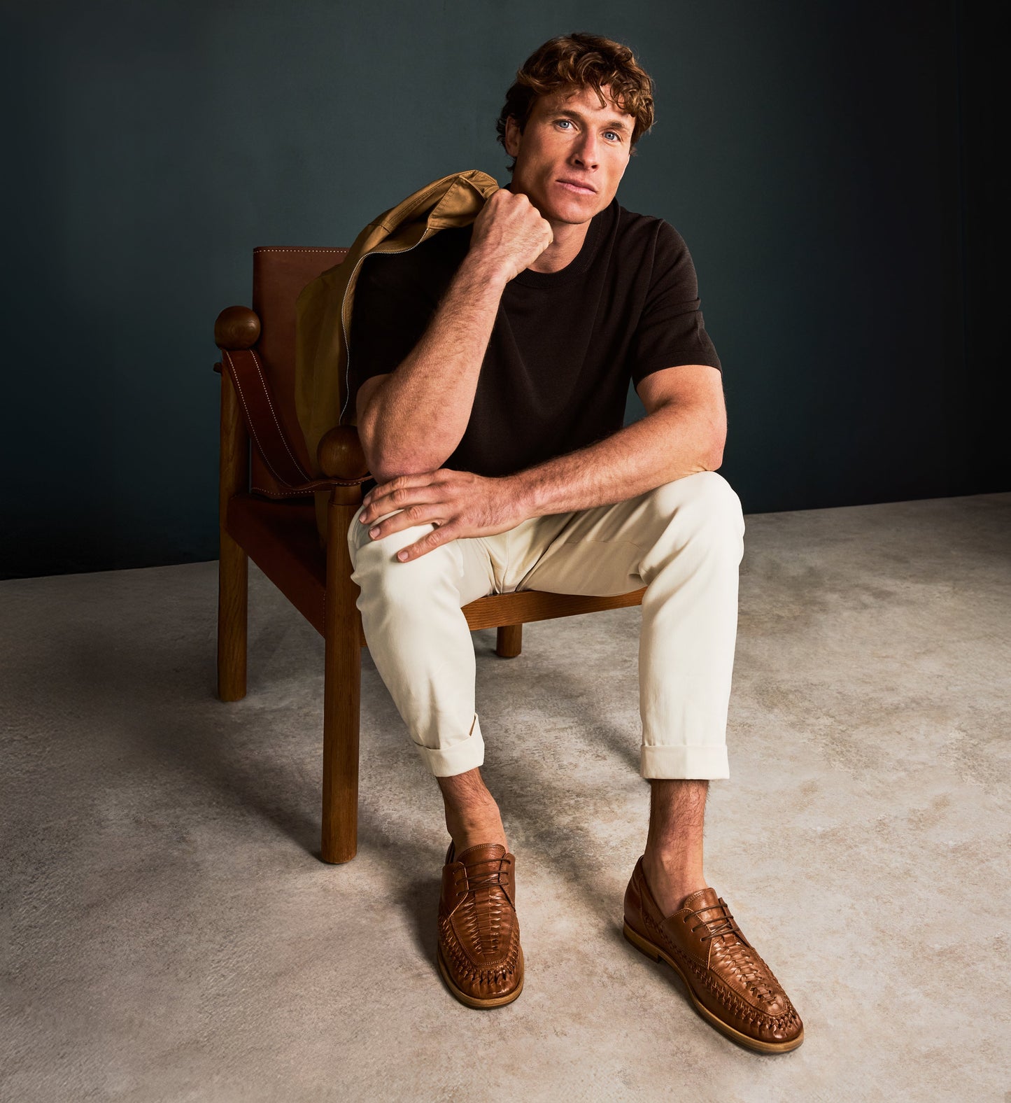 Man sitting on a wooden chair wearing a black shirt, beige pants, and brown shoes against a dark background.