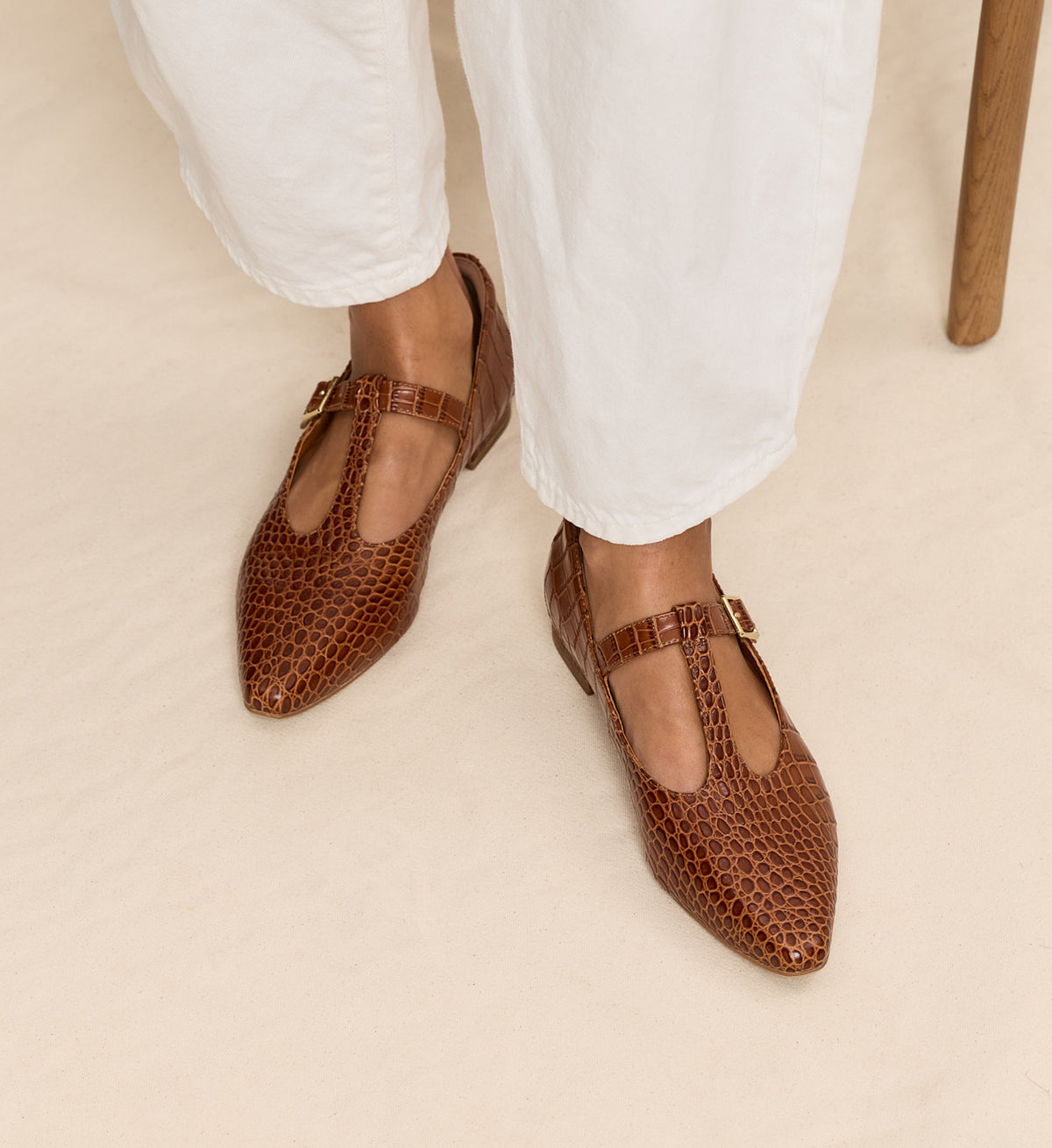 Brown pointed-toe flats worn with white pants on a beige background