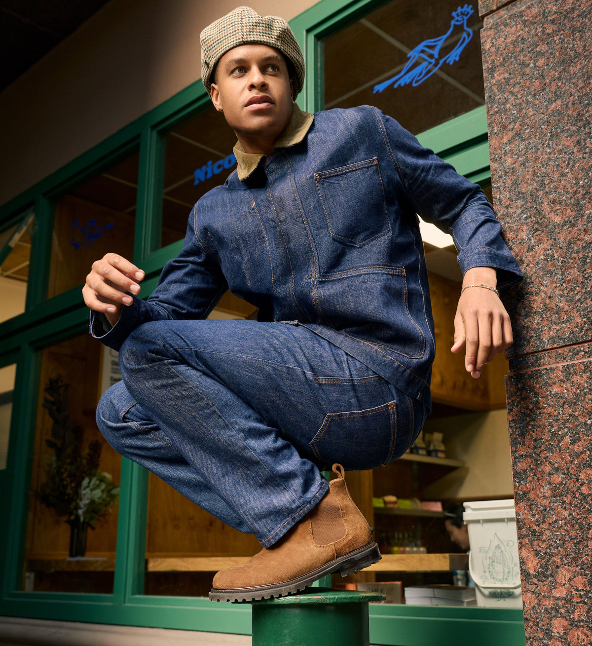 Person wearing a blue denim jumpsuit and tan suede chelsea boots leaning against a building.