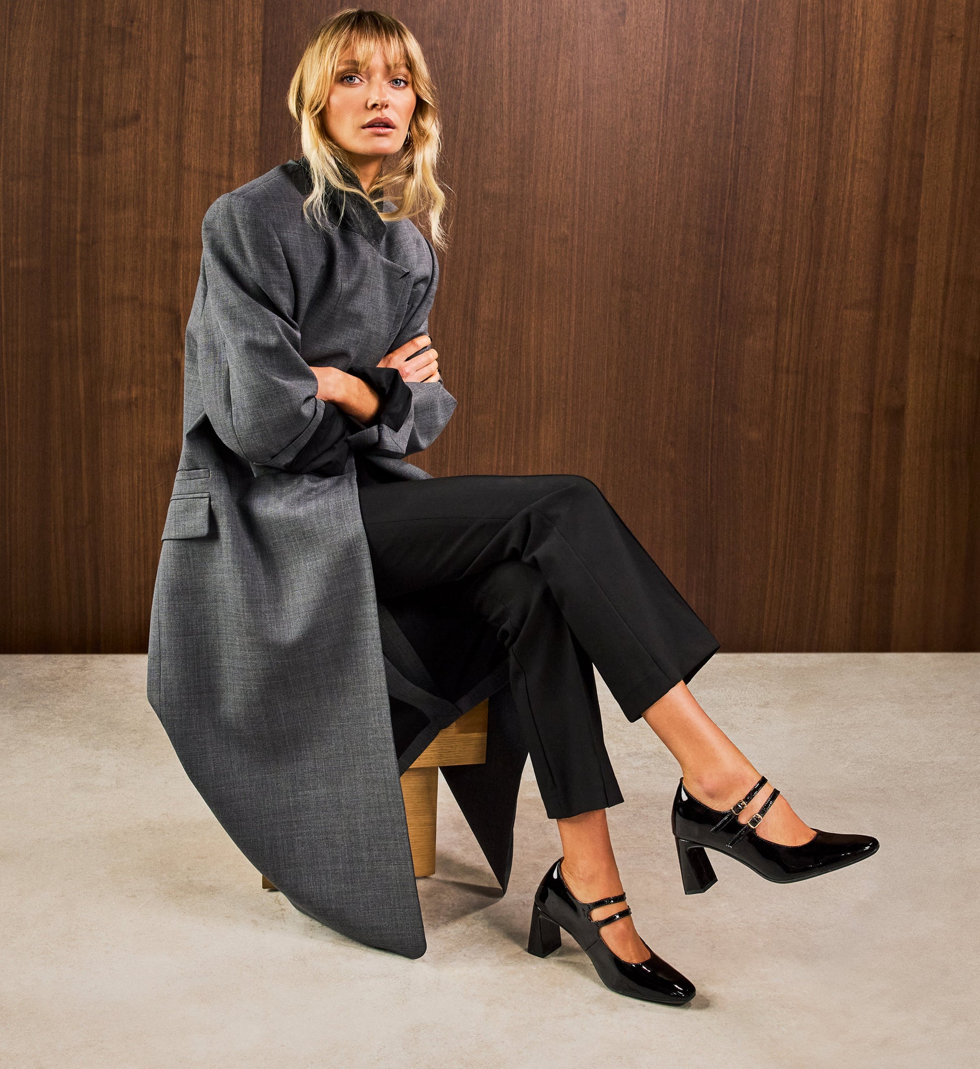 Woman wearing a long gray coat and black pants sitting on a wooden stool against a wooden wall with bared footwear patent closed toe black heels