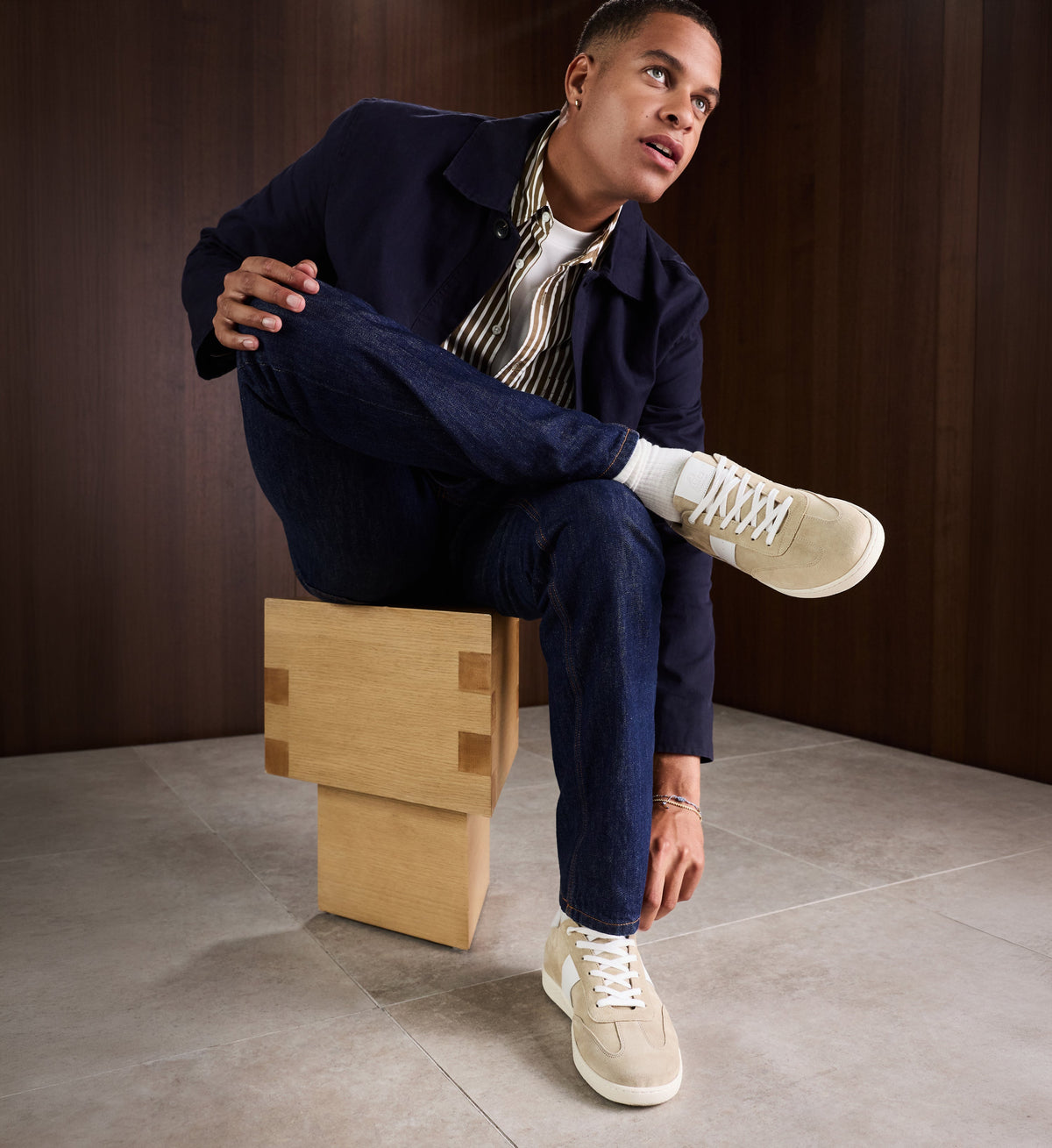 Man sitting on a wooden stool wearing a navy coat, striped shirt, and beige suede sneakers.