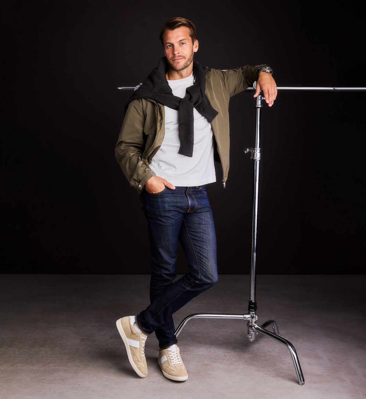 Man posing in front of a camera stand against a black background with beige suede sneakers, jeans and a green jacket