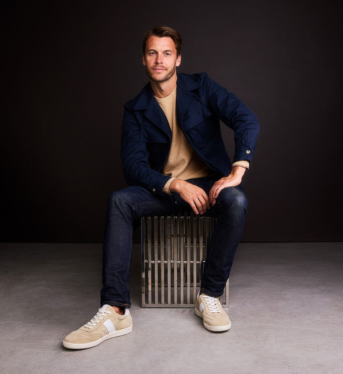 Man sitting on a metal crate wearing a navy jacket, beige shirt, and beige sneakers against a black background.