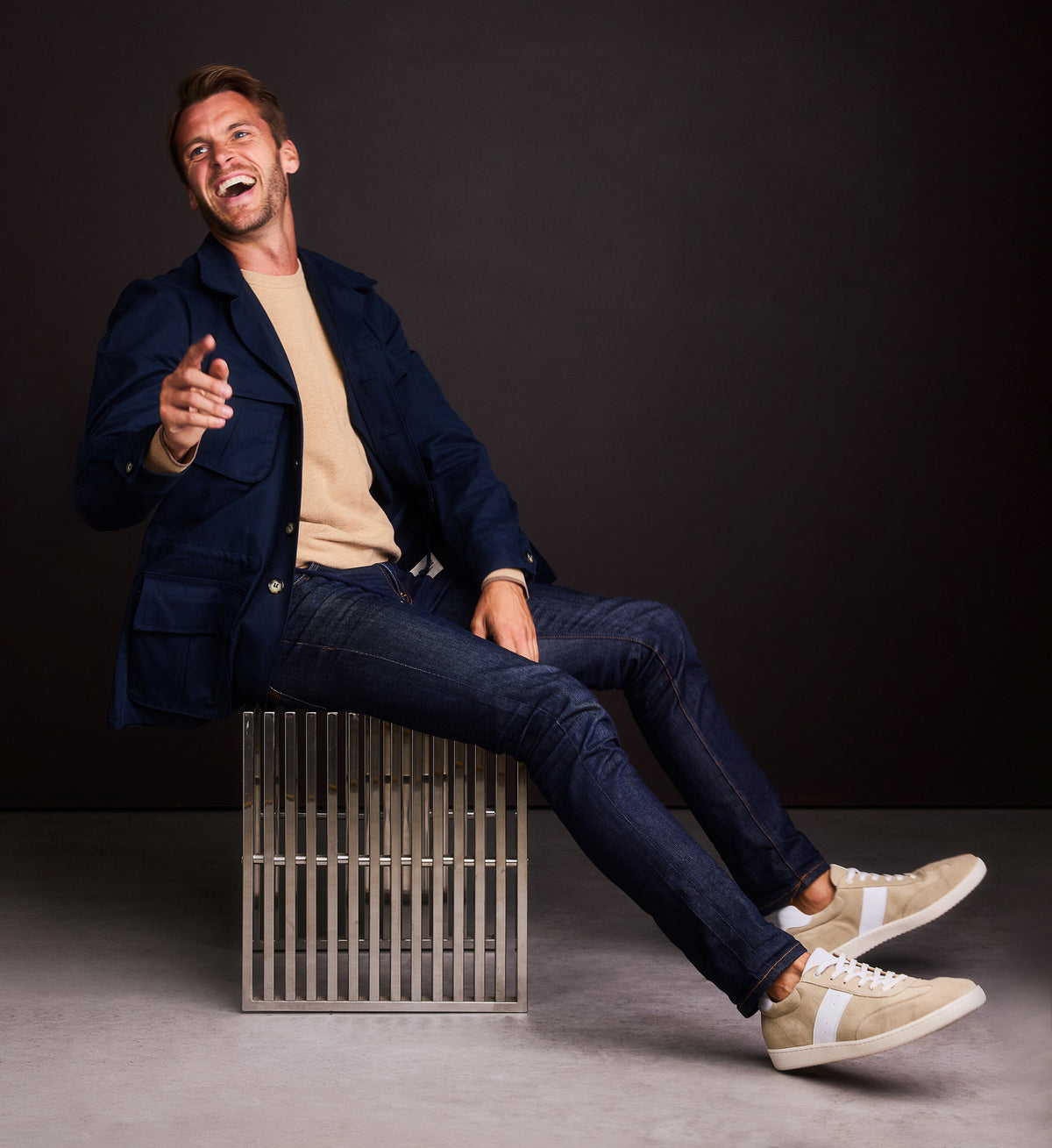 Man sitting on a metallic stool against a dark background wearing jeans and beige sneakers