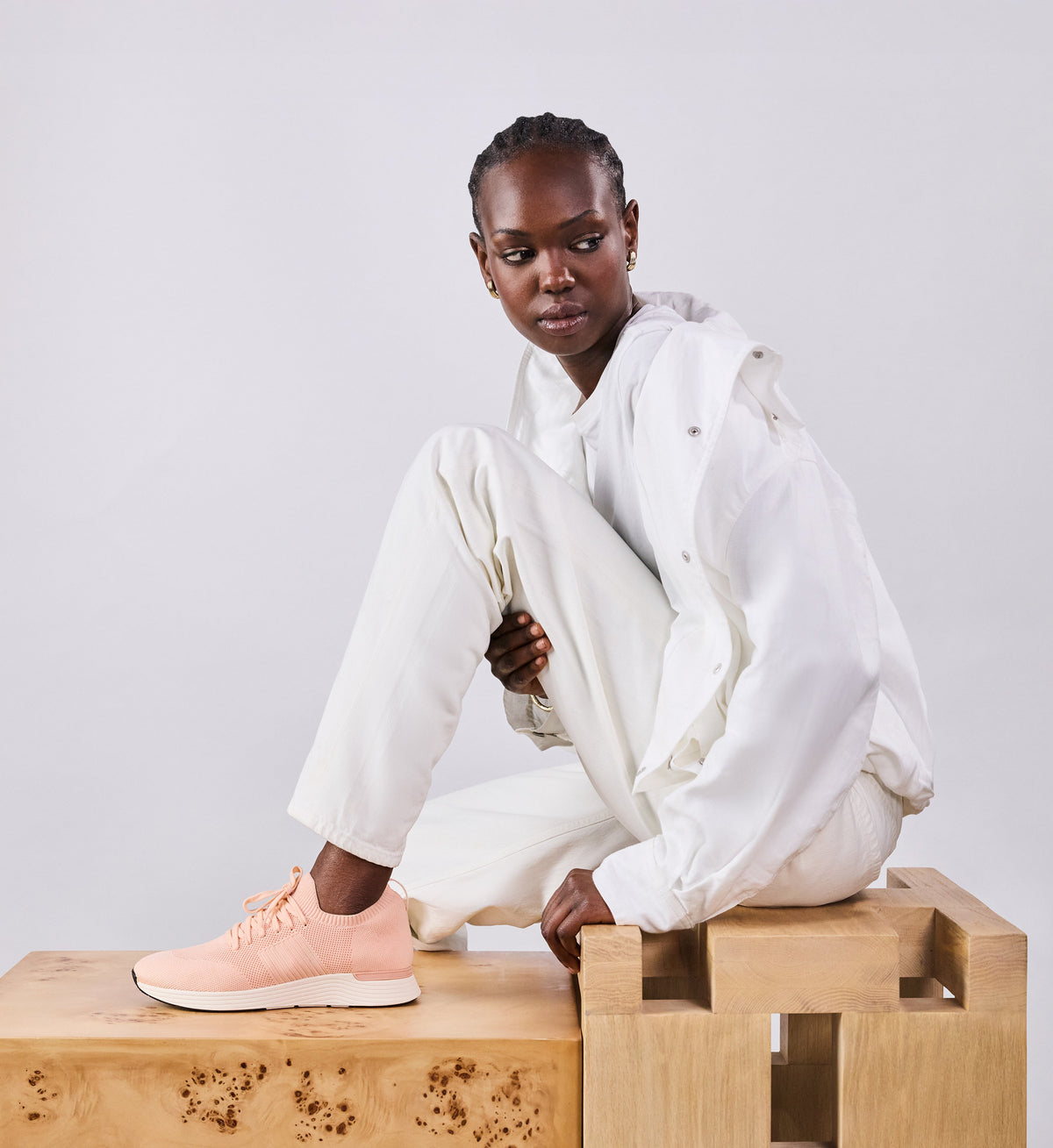 Person wearing a white outfit with pink shoes sitting on a wooden block against a light gray background