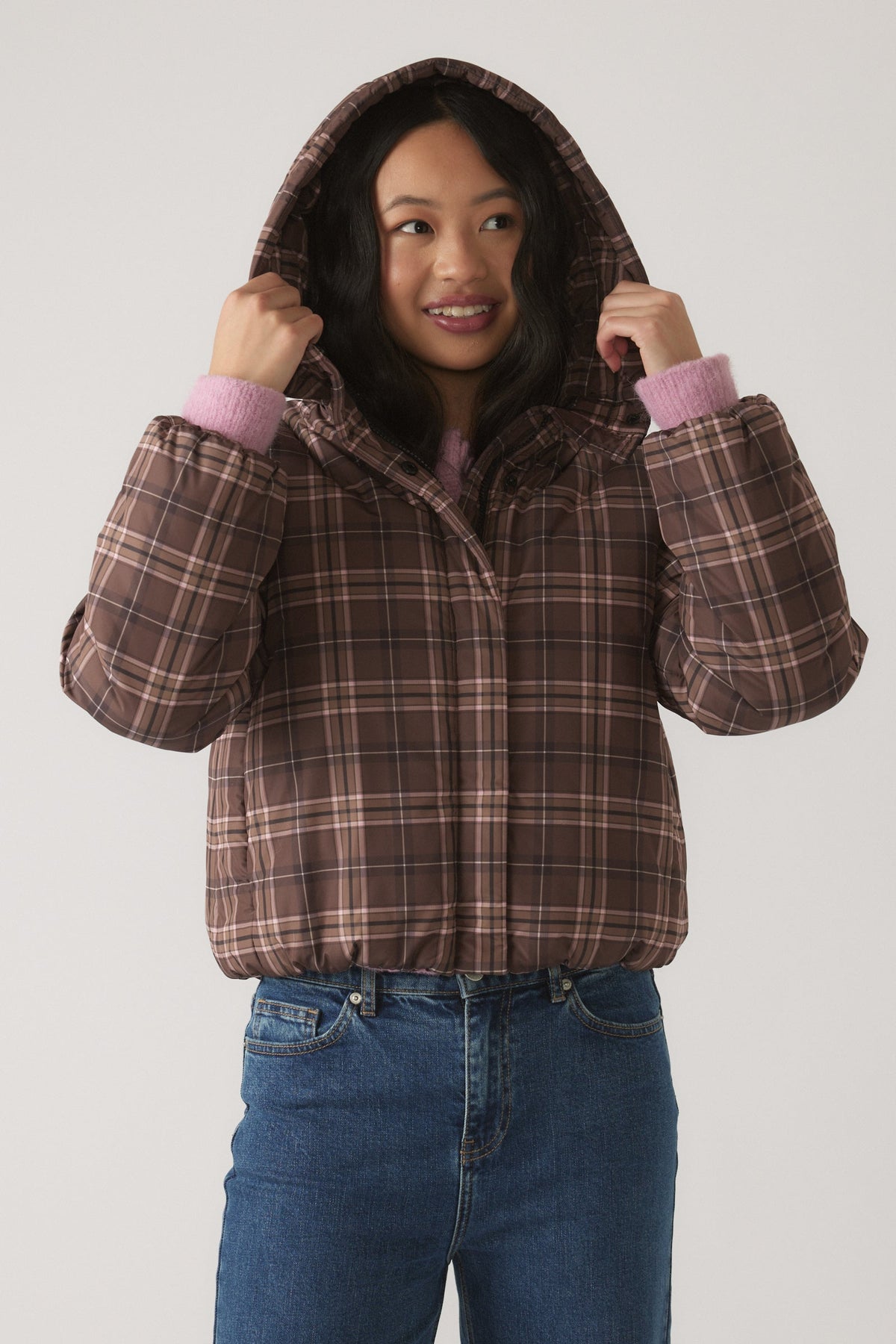 Girl wearing a cozy brown check hooded puffer jacket layered over a soft pink sweater and casual blue jeans