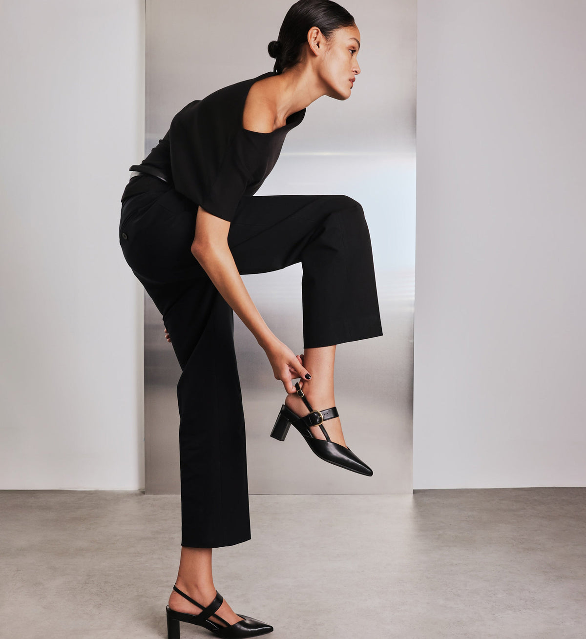 Woman in black outfit adjusting her black shoes against a plain background
