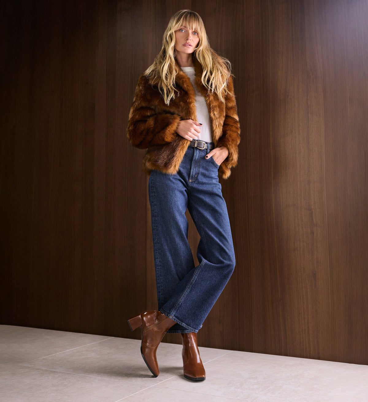Woman wearing a fur coat, jeans, and tan leather boots against a wooden wall.