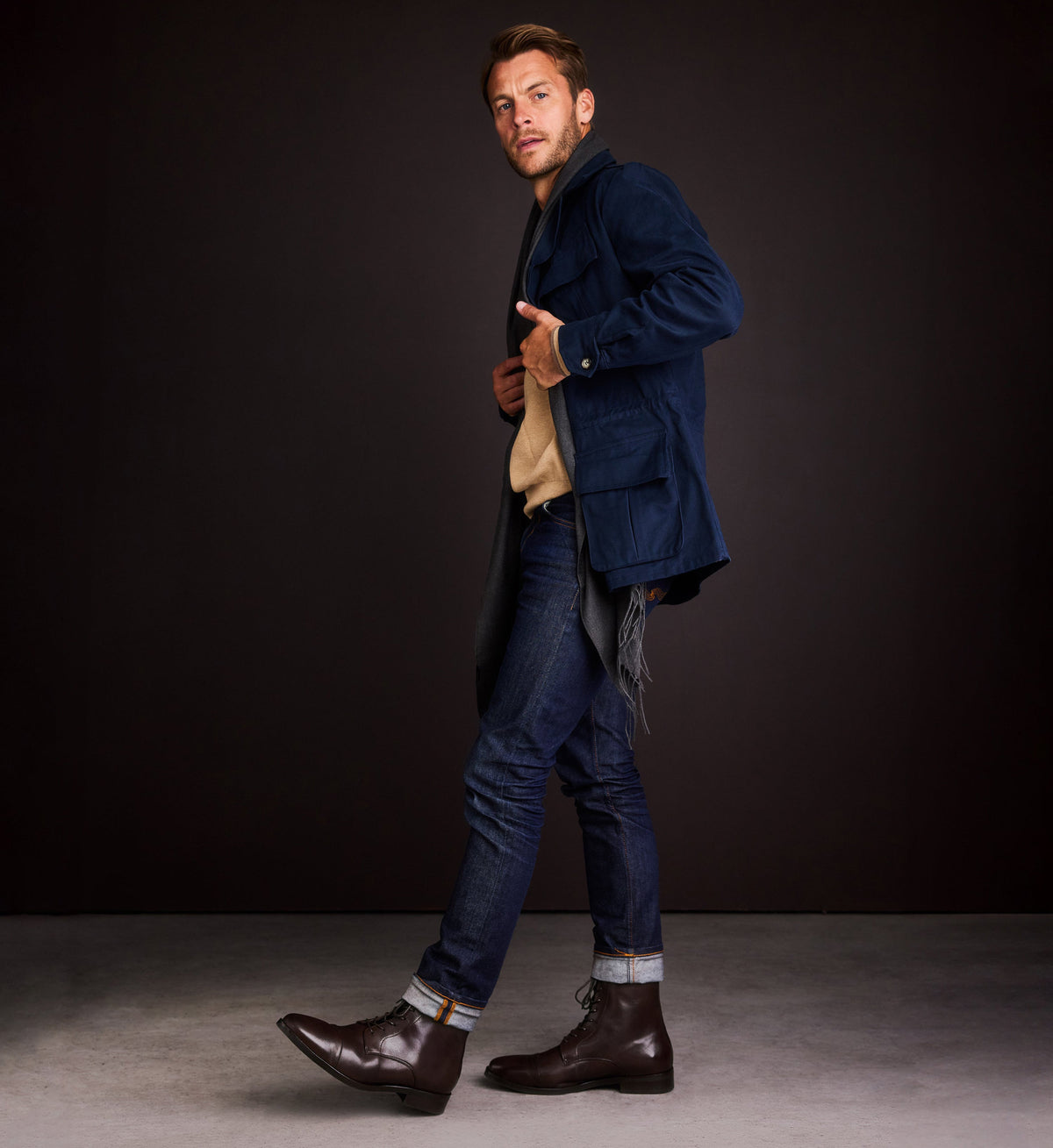 Man wearing a blue coat and jeans against a dark background with brown lace up boots