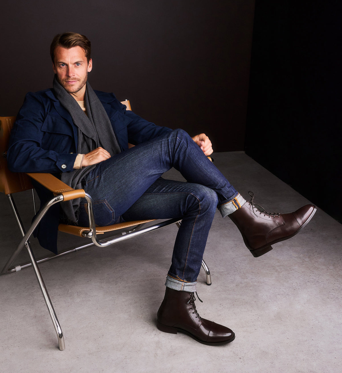 Man sitting on a chair wearing a navy coat, jeans, and brown boots.