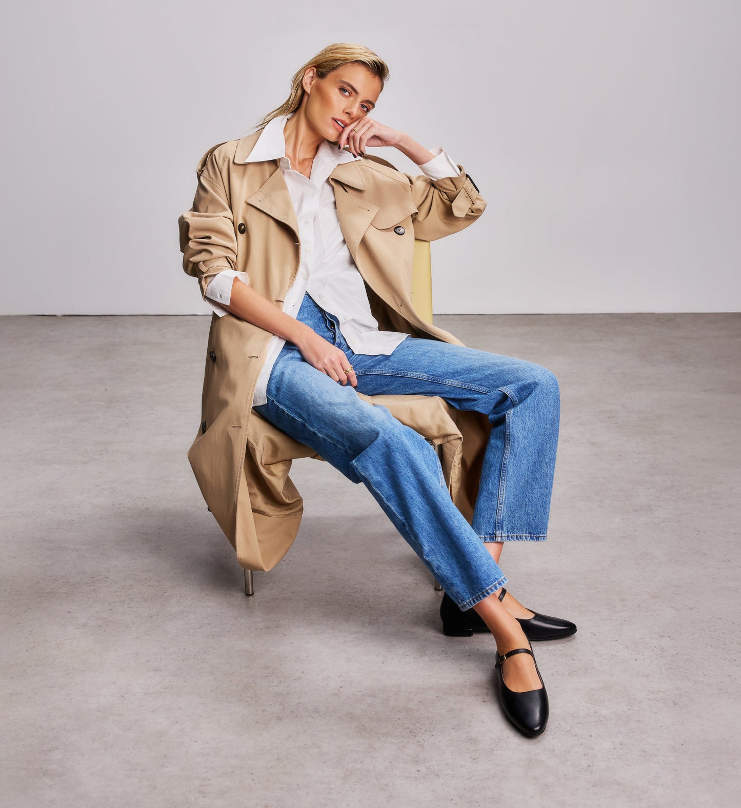 woman sitting on a chair wearing a trench coat, blue jeans and black ballet flats