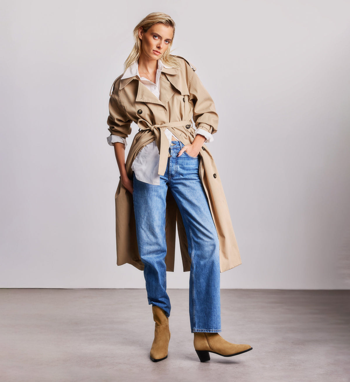 Woman wearing a beige trench coat, blue jeans, and tan suede boots on a plain background