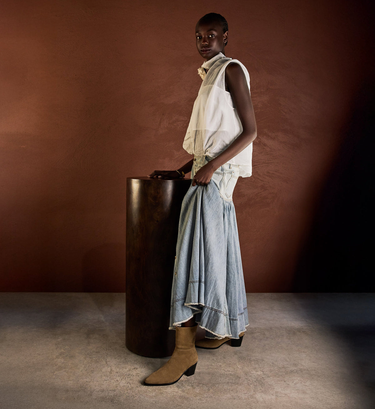 Person wearing a white sleeveless top and blue denim skirt standing next to a wooden cylindrical object against a brown background with tan suede boots