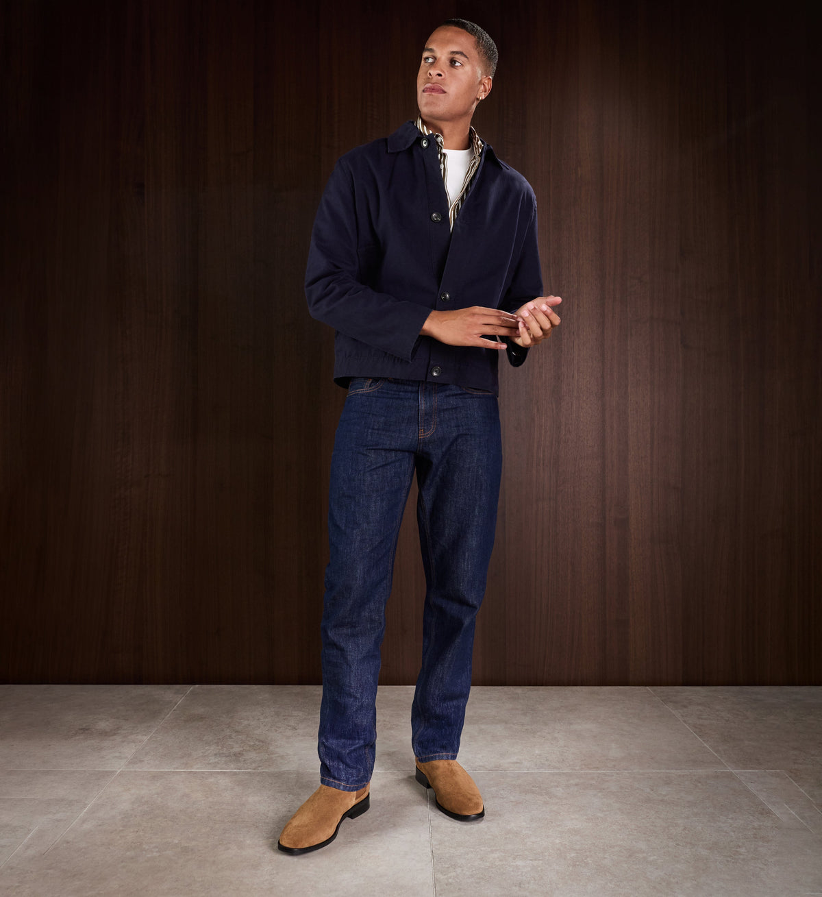 Man wearing a navy jacket, white shirt, blue jeans, and tan suede boots against a dark wooden background.