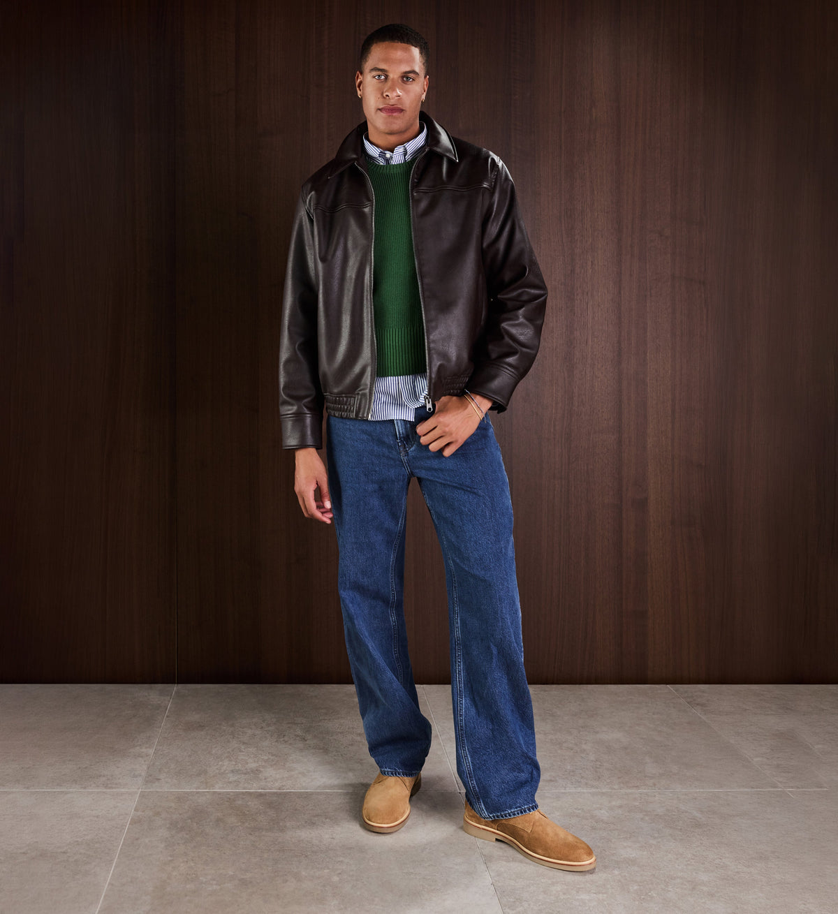 Man wearing a leather jacket, green sweater, blue jeans, and tan suede shoes against a wooden background.