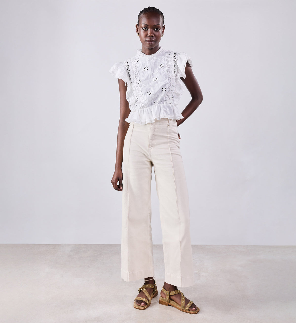 Woman wearing a white embroidered top and beige pants against a plain background with suede sandals with studs