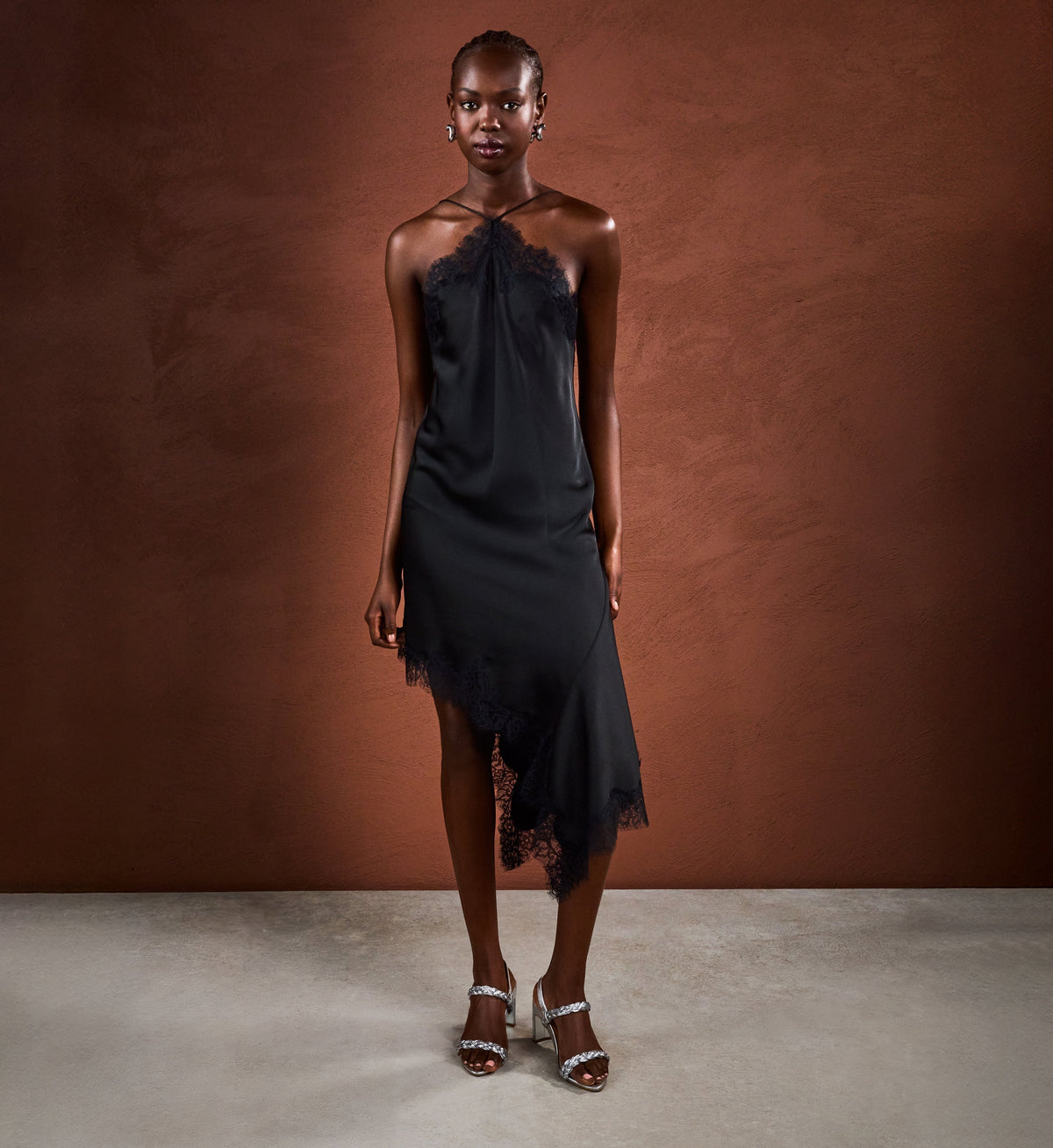Woman wearing a black halter dress against a brown background with silver heels with woven straps