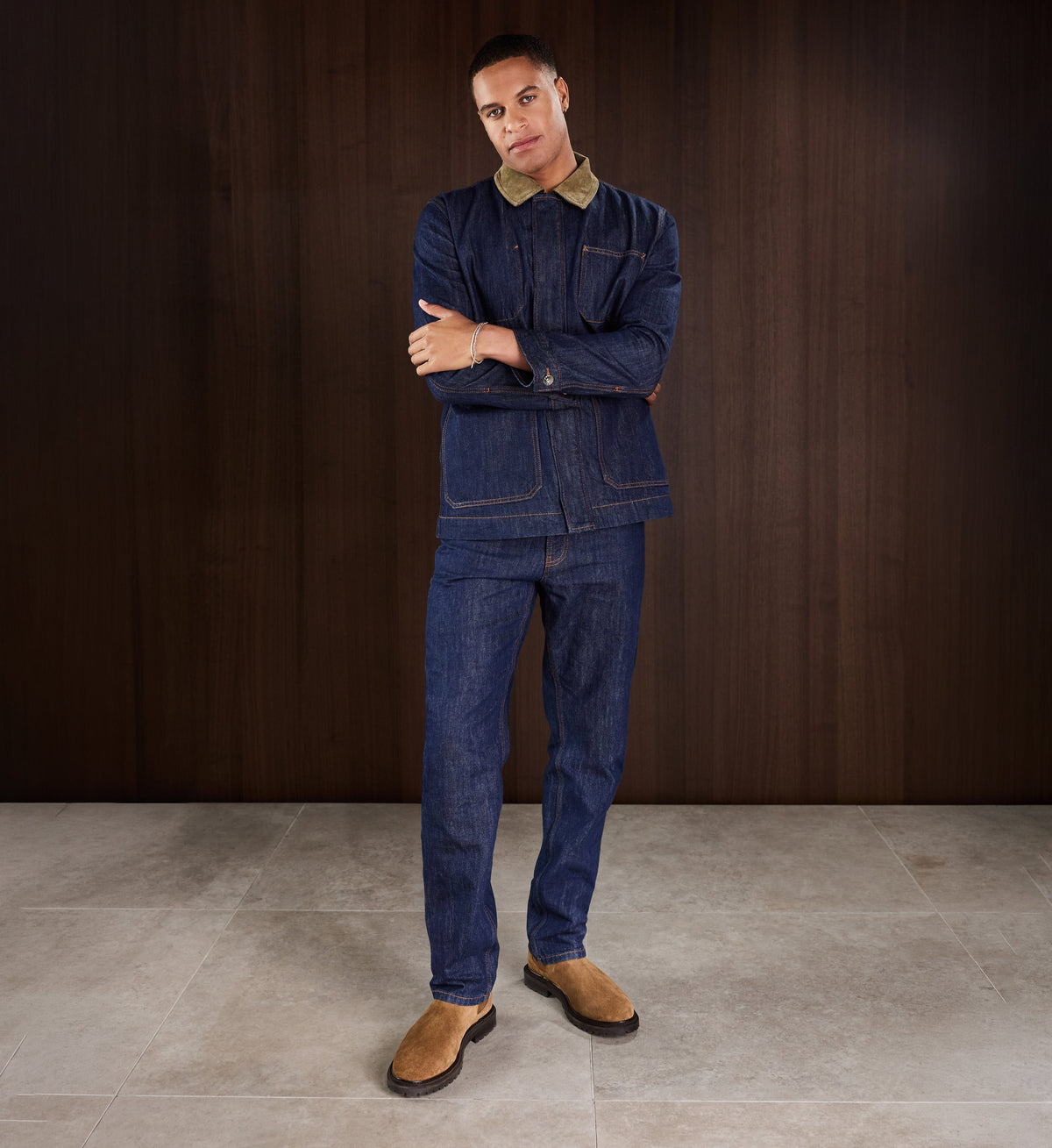 Man wearing a blue denim outfit with a brown shirt underneath, standing against a dark wooden wall wearing suede boots