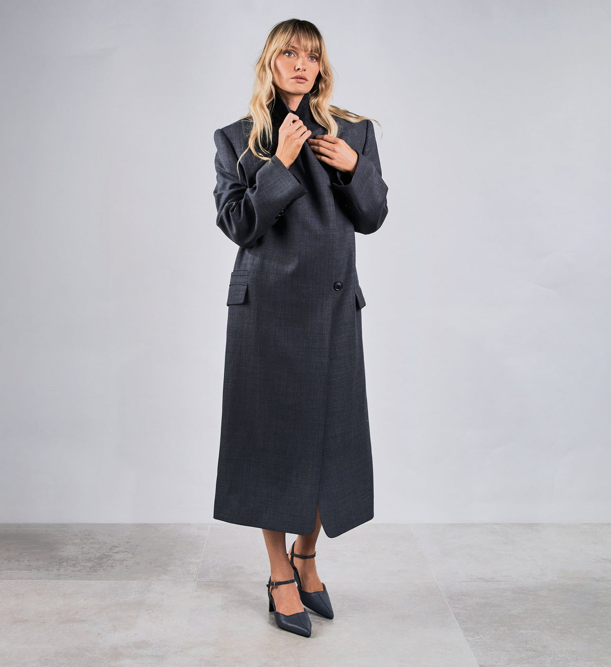 Woman wearing a long dark coat against a plain background wearing grey pointed toe heels