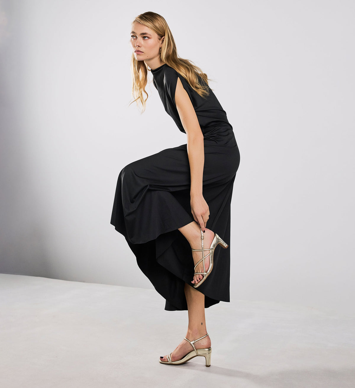 Woman in a black dress with gold heels posing on a light gray background