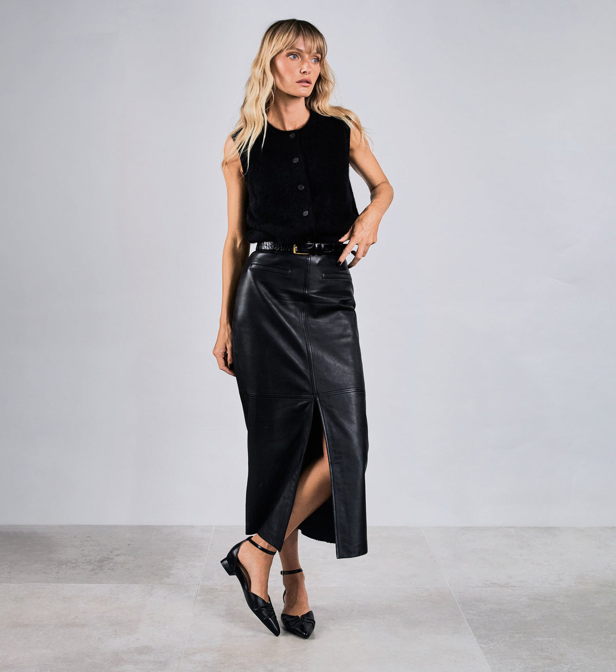 Woman wearing a black sleeveless top and leather skirt with black flats against a plain background