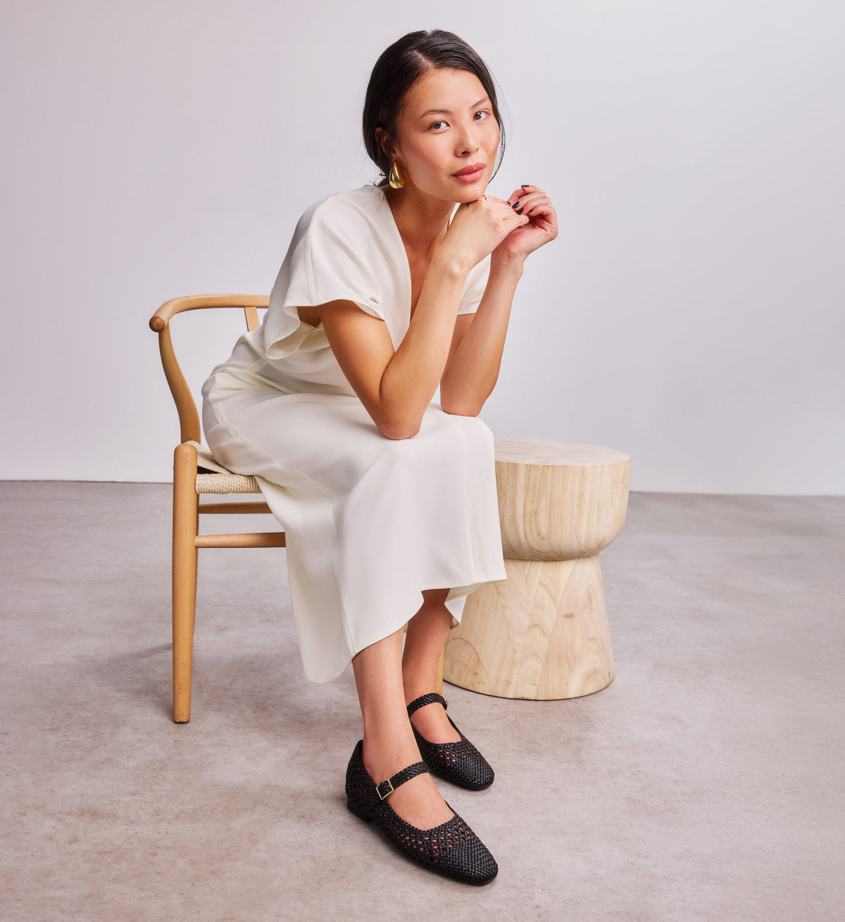 Woman in a white dress sitting on a wooden chair with a neutral background with black woven ballet flats