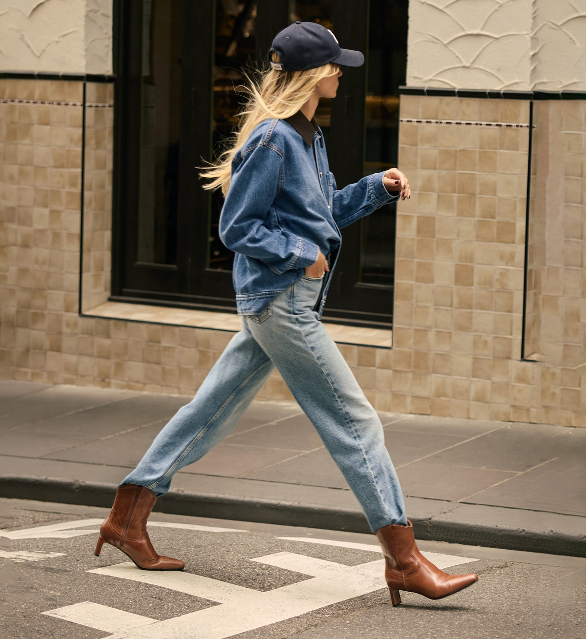 Woman walking on a street wearing a blue denim jacket, light blue jeans, and tan cowboy boots.
